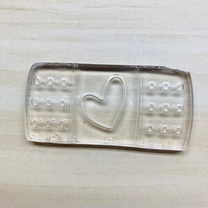 以下が含まれることがあります： ハート型の切り抜きのある透明なプラスチック製のばんそうこう。ばんそうこうは、小さな穴のあるテクスチャ加工された表面をしています。