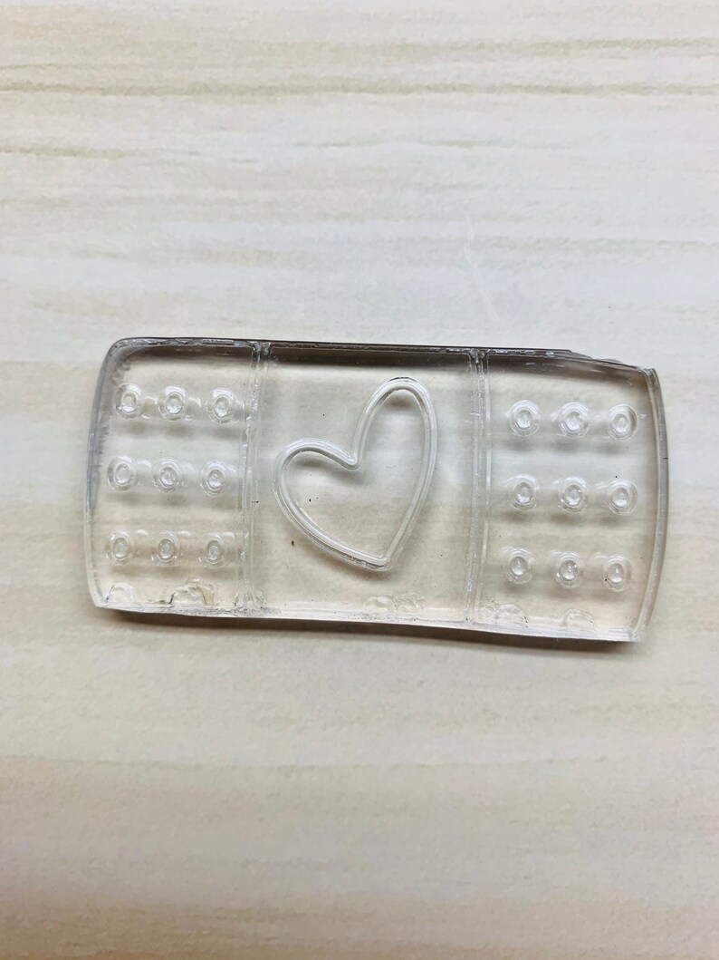 以下が含まれることがあります： ハート型の切り抜きのある透明なプラスチック製のばんそうこう。ばんそうこうは、小さな穴のあるテクスチャ加工された表面をしています。