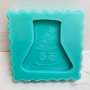 May include: A teal blue silicone mould shaped like a beaker with a smiling face and two eyes. The mould is designed for making resin crafts.