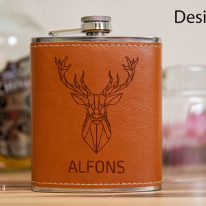 Könnte beinhalten: Eine braune, lederbezogene Trinkflasche mit silbernem Verschluss und einem gravierten geometrischen Hirschmotiv auf der Vorderseite. Die Trinkflasche ist mit dem Namen "ALFONS" personalisiert.