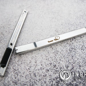 Könnte beinhalten: Ein silbernes Cuttermesser mit schwarzem Griff und einer personalisierten Gravur mit der Aufschrift "Bester Opa" und einem Herzsymbol.