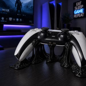 Op de afbeelding: Een zwart-witte gamecontroller op een zwarte, gestileerde standaard. De standaard heeft een uniek, gesmolten ontwerp. De controller staat centraal, met een vage achtergrond met een monitor met een videogame en een bord met de tekst "EAT SLEEP GAME REPEAT".