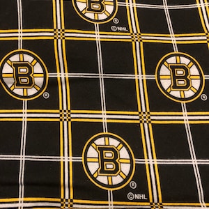 May include: Black and yellow fabric with a repeating pattern of the Boston Bruins logo. The logo is a black "B" inside a circle with white and yellow stripes. The fabric also has a black and white plaid pattern. The text "NHL" is printed in white on the fabric.