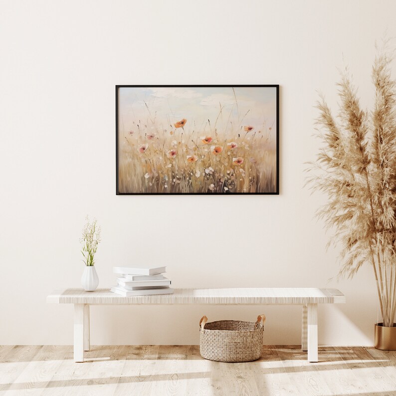 May include: A framed painting of a field of wildflowers with a light blue sky in the background. The painting is hanging on a white wall above a white bench with a woven basket and a vase of white flowers.