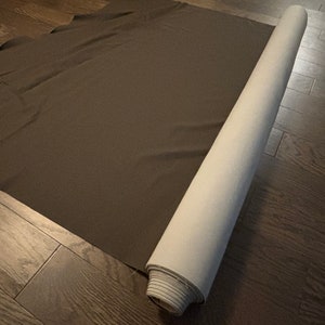 May include: A roll of dark gray fabric next to a flat piece of the same material. The fabric is on a dark wood floor. The rolled fabric is white.