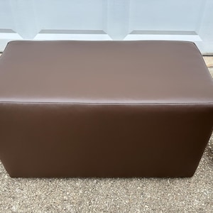 May include: A brown leather ottoman with a rectangular shape. The ottoman is sitting on a gravel surface.