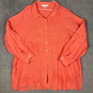 May include: A long-sleeved button-down shirt in a vibrant coral orange color. The shirt features a classic collar, a full-length front button placket, and buttoned cuffs. The fabric has a visible textured weave, and a small white label is at the neck.