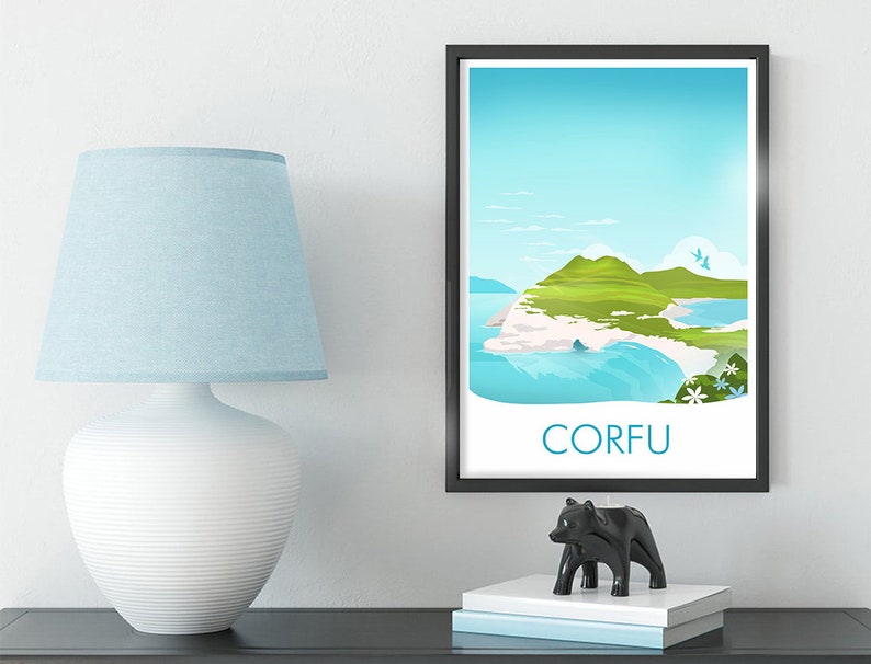 May include: A framed art print of Corfu island with a black frame. The print features a coastal landscape with green hills, blue water, and the word "CORFU" in white. A white lamp with a blue lampshade and a black bear figurine are on a dark surface.