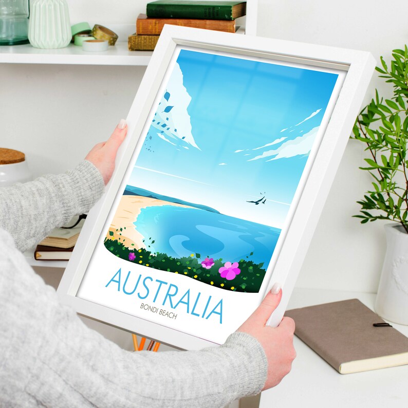 May include: A white framed print featuring a colourful illustration of Bondi Beach in Australia. The print depicts a blue sky with white clouds, a blue ocean with white waves, and green trees and flowers. The text "Australia" and "Bondi Beach" is printed in blue on the print.