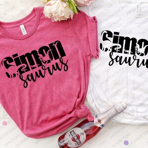 May include: Two pink and white t-shirts with black text that reads "Simon Saurus". The shirts are folded and laid out on a surface with a bottle of sparkling wine.