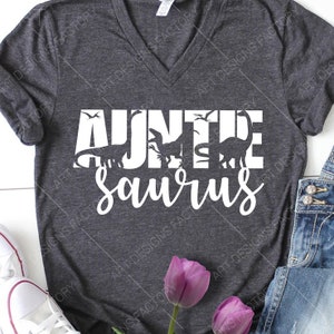 May include: A dark grey v-neck t-shirt featuring the white text "Auntie Saurus" with dinosaur silhouettes incorporated into the lettering. This casual t-shirt is suitable for everyday wear.