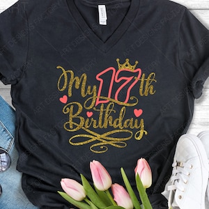 May include: A black t-shirt with a gold glitter design that says "My 17th Birthday" with a crown above the number 17 and two pink hearts on either side of the text.