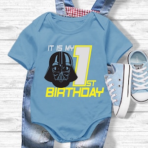 May include: A light blue baby onesie with a black Darth Vader graphic and the text "It is my 1st Birthday" in yellow. The onesie is paired with light blue denim overalls and light blue sneakers.
