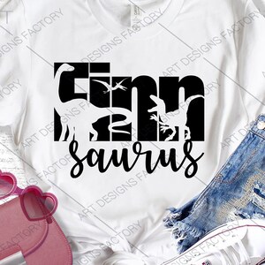 May include: White t-shirt with a black graphic design that says "Tim Saurus" with dinosaur silhouettes. The graphic is made up of the letters of the name.