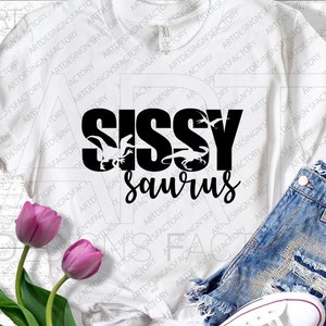 May include: White t-shirt with black text that reads "Sissy Saurus" with a dinosaur graphic. The shirt is laying on a wooden surface with a pair of denim shorts and a pair of white sneakers.