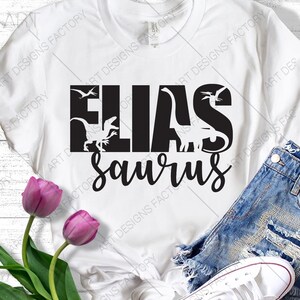 May include: White t-shirt with the name "Elias" in black block letters, and "Saurus" in cursive script below. Dinosaur silhouettes are integrated into the design. The image includes denim shorts, white trainers, and purple tulips, creating a casual aesthetic.