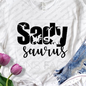 May include: White t-shirt with black text that reads "Sadly Saurus". The text is in a playful font and features a dinosaur silhouette.