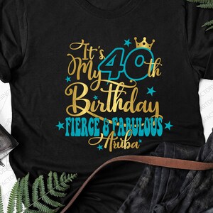 Può includere: Maglietta nera con testo dorato e blu che recita "It's My 40th Birthday Fierce & Fabulous Aruba". Il numero 40 è in un cerchio blu con una corona dorata sopra.