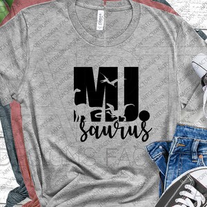 May include: Grey t-shirt with a black graphic design that says "MJsaurus". The graphic features a dinosaur silhouette and a bird.