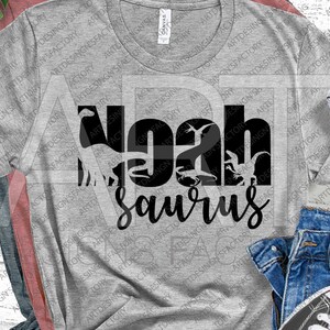May include: A grey t-shirt with a black graphic design that says "Noah Saurus" with dinosaur silhouettes incorporated into the letters.