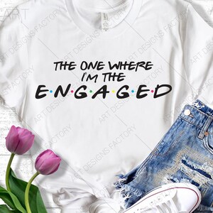 Puede incluir: Camiseta blanca con el texto "The One Where I'm The Engaged" en negro con puntos de colores. El texto está estilizado como la popular serie de televisión "Friends".