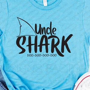 May include: A light blue t-shirt featuring a black shark fin and the text "Uncle Shark Doo-Doo-Doo-Doo" in black.