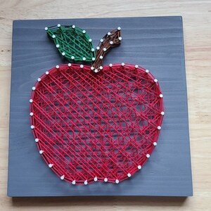 May include: A red apple with a green leaf and brown stem is created with string wrapped around nails on a gray wooden board.
