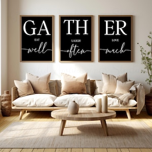 May include: Three framed wall art pieces with the words "GATHER, LAUGH, LOVE" in large white letters on a black background. Below each word are the words "EAT well", "LAUGH often", and "LOVE much" in a script font. The art hangs above a light-coloured sofa.