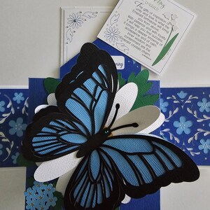 May include: A blue and black paper butterfly with a white background. The butterfly is surrounded by blue and white flowers. The image also includes a white card with the text "May Emerald The gifts of inspiration of unity and love is what the Emerald birthstone will bring you from above. With patience and with loyalty it will help you find success. brings a deeper knowing of acceptance and of calm. Inner wisdom and contentment are the Emerald crystals charm. ning"