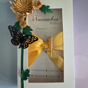 May include: A white card with a window showing a yellow bow, a black butterfly, and green leaves. The card says "November Birthday Gemstone Topaz" and "Faithfulness, Constancy, Friendship, Loyalty".