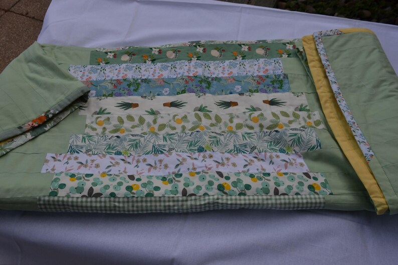 May include: A handmade quilt with a patchwork design featuring various floral and botanical patterns in shades of green, white, and blue. The quilt has a light green border and a yellow binding. The quilt is made of fabric.