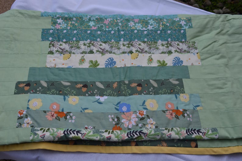 May include: A handmade quilt with a patchwork design. The quilt features horizontal stripes of various floral and leaf patterns in shades of green, blue, and cream. The edges are a solid light green, and the quilt is approximately 60 inches wide and 80 inches long.