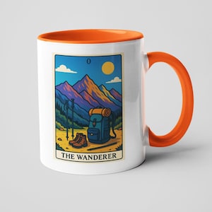 May include: White ceramic mug with an orange interior and handle. The mug features a tarot card design with a mountain landscape, backpack, hiking boots, and the text "THE WANDERER".
