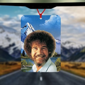 May include: A car air freshener featuring a portrait of a man with a bushy afro, smiling against a backdrop of a mountain range and a blue sky.