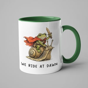May include: White ceramic mug with a green interior and handle. The mug features a whimsical illustration of a frog riding a snail, holding a sword, and wearing a red cape. The text "WE RIDE AT DAWN" is printed on the mug.