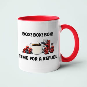 May include: A white ceramic mug with a red interior and handle. The front features a graphic of small figures in red racing suits surrounding a coffee cup, with the text "BOX! BOX! BOX!" above and "TIME FOR A REFUEL" below the illustration.