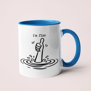 May include: A white ceramic mug with a blue interior and handle. The exterior features a black line drawing of an arm emerging from water with a thumb up gesture and the text i'm fine printed in a typewriter font above the illustration.
