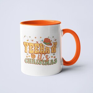 May include: A white ceramic mug with an orange interior and handle. The front features a graphic with the text YEEHAW it's CHRISTMAS in yellow and green letters, a brown cowboy hat with a white trim, a disco ball, and colorful string lights.