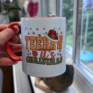 May include: A white ceramic mug with an orange handle and interior. The front features the text YEEHAW it's CHRISTMAS in orange and green lettering, decorated with a cowboy hat, colorful string lights, and small star shapes.