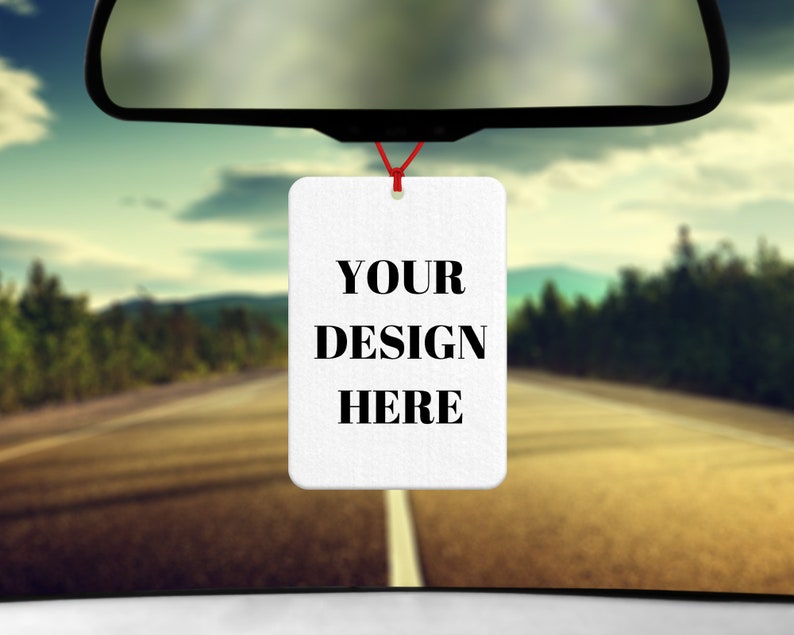 Puede incluir: Un ambientador de coche blanco con el texto "YOUR DESIGN HERE" colgado del espejo retrovisor de un coche. El coche est&aacute; conduciendo por una carretera con &aacute;rboles a ambos lados.