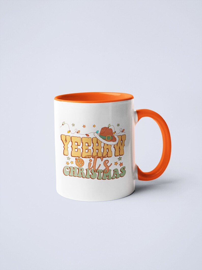May include: A white ceramic mug with an orange interior and handle. The front features a graphic with the text YEEHAW it's CHRISTMAS in yellow and green letters, a brown cowboy hat with a white trim, a disco ball, and colorful string lights.