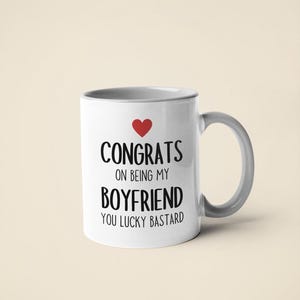 A white ceramic mug with black text that reads "CONGRATS ON BEING MY BOYFRIEND YOU LUCKY BASTARD" and a red heart symbol.