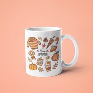 May include: White ceramic mug featuring autumn-themed illustrations in orange and brown, including a sweater, leaves, pumpkin, and the text "Hello Autumn." The mug has a curved handle and is set against a peach background.