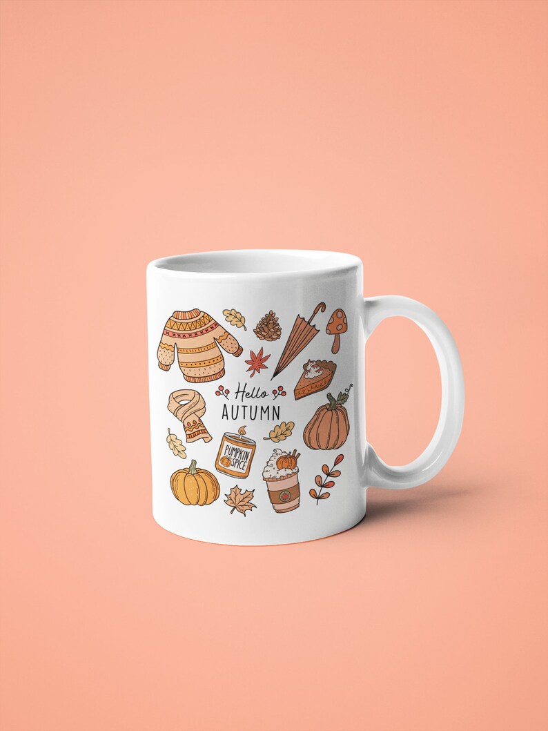 May include: White ceramic mug featuring autumn-themed illustrations in orange and brown, including a sweater, leaves, pumpkin, and the text "Hello Autumn." The mug has a curved handle and is set against a peach background.