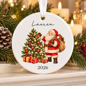 May include: A round white ceramic ornament featuring a graphic of Santa Claus decorating a Christmas tree with red and gold ornaments. The name Lauren is printed in black script at the top, and the year 2026 is printed in black at the bottom.