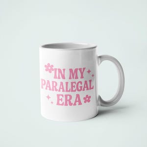 Op de afbeelding: Witte keramische mok met een gebogen handvat. De mok heeft de tekst "IN MY PARALEGAL ERA" in roze, gestileerde tekst met bloem- en steraccenten. De mok staat tegen een effen witte achtergrond.
