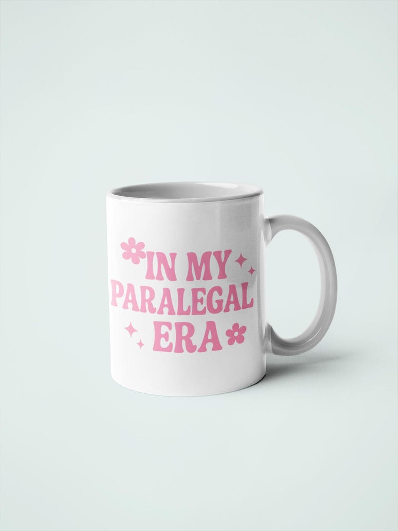 Op de afbeelding: Witte keramische mok met een gebogen handvat. De mok heeft de tekst "IN MY PARALEGAL ERA" in roze, gestileerde tekst met bloem- en steraccenten. De mok staat tegen een effen witte achtergrond.