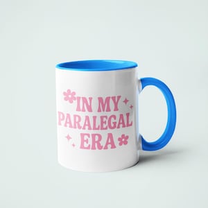 Op de afbeelding: Witte keramische mok met een blauwe binnenkant en handvat. De mok heeft de roze tekst "IN MY PARALEGAL ERA" met bloemen- en sterrenafbeeldingen. De mok staat tegen een effen witte achtergrond.