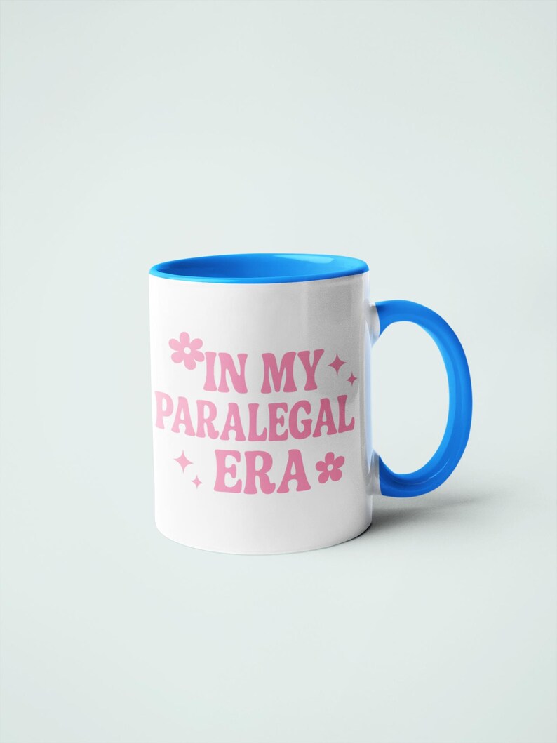 Op de afbeelding: Witte keramische mok met een blauwe binnenkant en handvat. De mok heeft de roze tekst "IN MY PARALEGAL ERA" met bloemen- en sterrenafbeeldingen. De mok staat tegen een effen witte achtergrond.