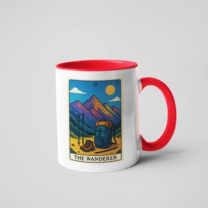 May include: White ceramic mug with a red interior and handle. The mug features a colorful illustration of a mountain scene with hiking gear and the text "THE WANDERER". The design includes a backpack, boots, and hiking poles.